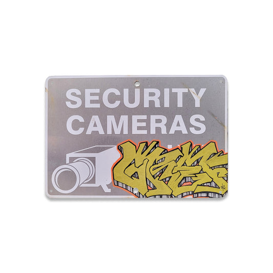 Metal security camera sign with yellow graffiti writing